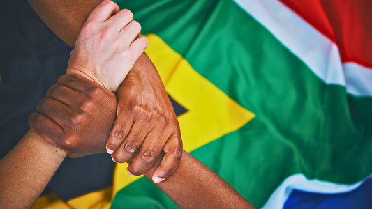 Three people of different races holding hands in front of the South African flag showing unity and the power of one nation.