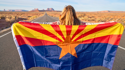 Woman standing in the middle of a long stretch of road holding an Arizona State Flag across her shoulders so that it can wave in the wind.