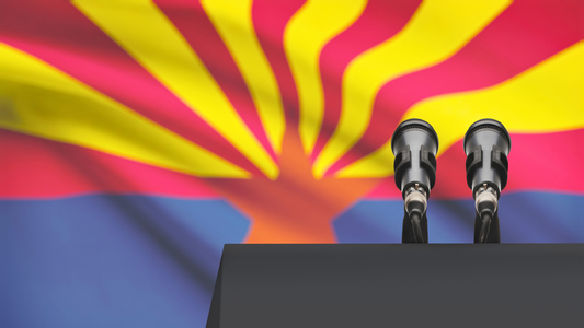 A partial view of a podium with two microphones. The Arizona State flag is hanging as a backdrop behind the podium.
