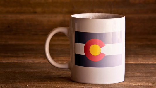 Coffee mug with a Colorado state flag printed on it. The mug is displayed on a wooden table.