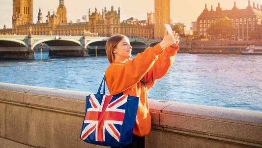 Travelling woman standing on a bridge taking a selfie. She as a Union Jack flag bag over her shoulder.