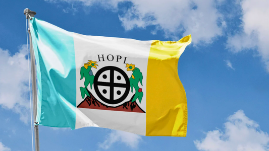The Hopi Nation flag showing rich symbolism, flying on a flagpole in front of a blue sky and scattered white clouds.