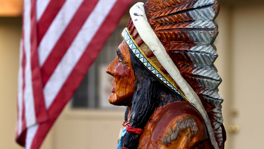 A Close-up of a wooden carving of a Native American Indian head with a part of a hanging US Flag visible in the background