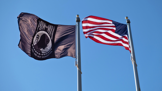 The POW/MIA flag and the US Flag flying next to each other, flapping in the wind with clear blue skies behind it.