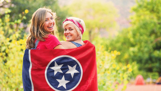 The TN Flag draped across the shoulders of two woman stading side by side looking back and smiling at the camera.